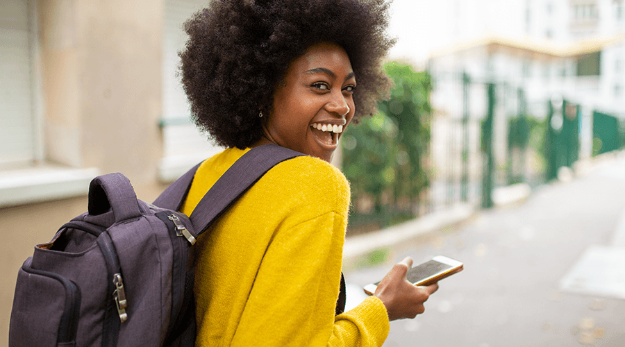 Female student smiling walking down the street wearing a backpack and holding a smartphone.